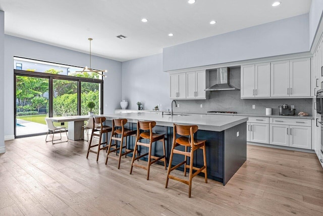 kitchen with pendant lighting, light wood-type flooring, a center island with sink, and wall chimney range hood