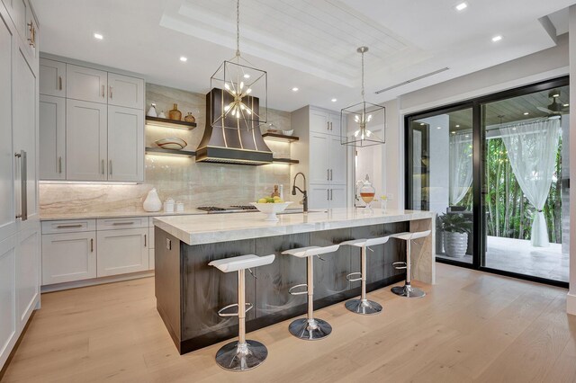 kitchen featuring backsplash, custom range hood, light hardwood / wood-style floors, a raised ceiling, and a center island with sink