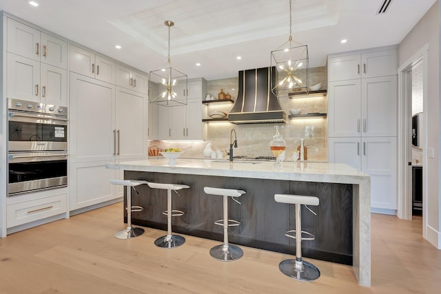 kitchen featuring wall chimney range hood, a tray ceiling, stainless steel double oven, and light hardwood / wood-style floors