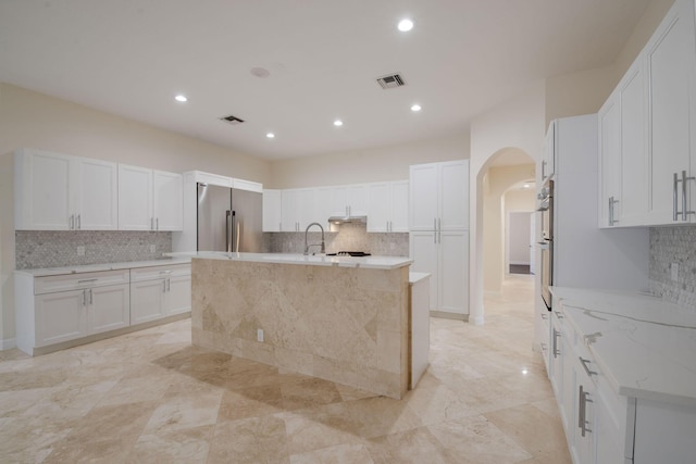 kitchen featuring white cabinetry, an island with sink, stainless steel refrigerator, light stone countertops, and sink