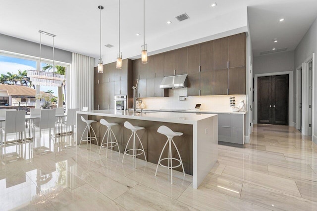 kitchen with a center island with sink, decorative light fixtures, ventilation hood, dark brown cabinets, and oven