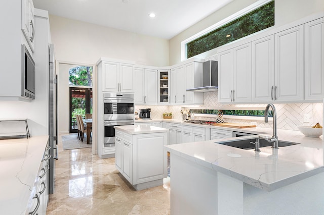 kitchen featuring wall chimney range hood, sink, appliances with stainless steel finishes, a kitchen island, and light stone counters