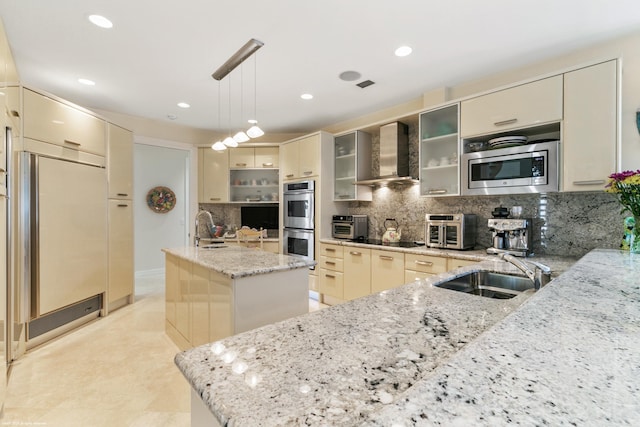 kitchen featuring wall chimney range hood, backsplash, an island with sink, built in appliances, and sink