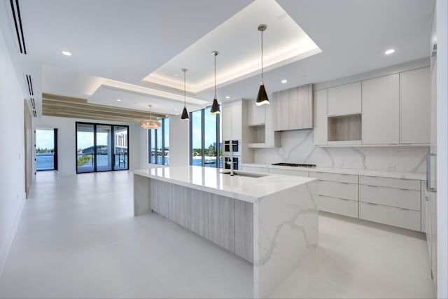 kitchen featuring decorative light fixtures, decorative backsplash, a center island with sink, and a raised ceiling