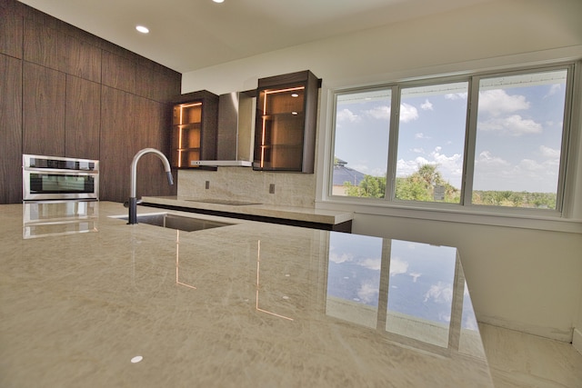 kitchen with decorative backsplash, wall chimney exhaust hood, oven, sink, and light stone counters