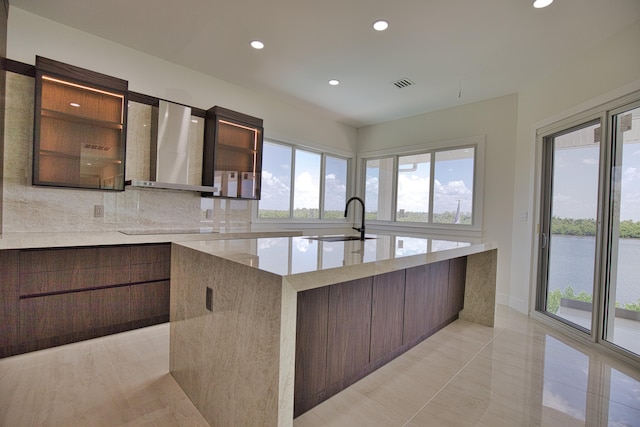 kitchen featuring wall chimney range hood, light tile patterned floors, a healthy amount of sunlight, and an island with sink