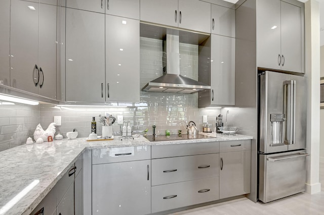 kitchen featuring high end refrigerator, wall chimney range hood, black electric stovetop, light stone countertops, and backsplash