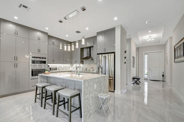 kitchen with gray cabinets, appliances with stainless steel finishes, an island with sink, a kitchen bar, and wall chimney range hood