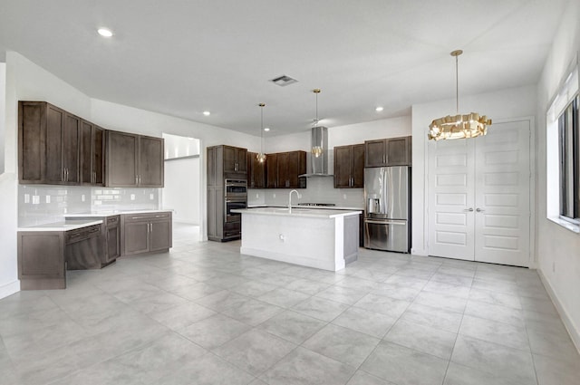 kitchen featuring decorative light fixtures, wall chimney range hood, stainless steel appliances, an island with sink, and light tile patterned floors