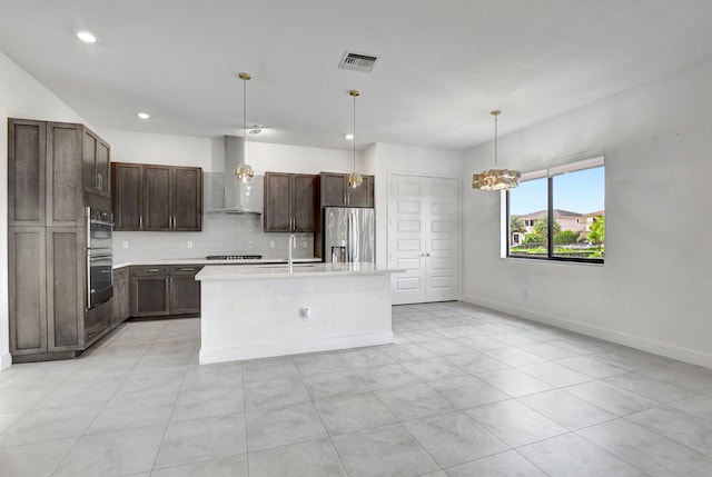 kitchen featuring decorative light fixtures, tasteful backsplash, wall chimney range hood, an island with sink, and appliances with stainless steel finishes