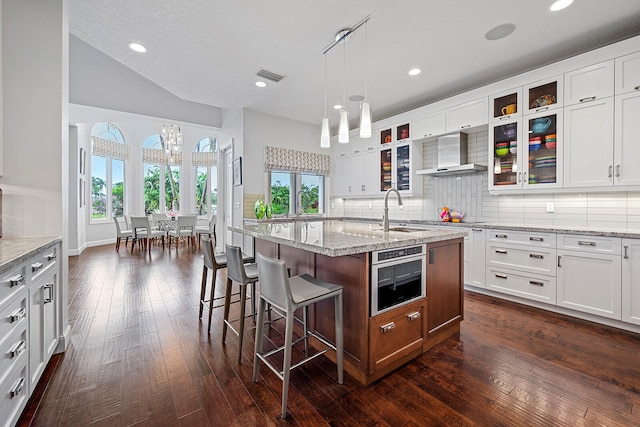 kitchen with decorative backsplash, wall chimney exhaust hood, oven, a center island with sink, and dark wood-type flooring