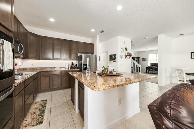 kitchen featuring light tile patterned flooring, dark brown cabinetry, light stone counters, stainless steel appliances, and a kitchen island with sink