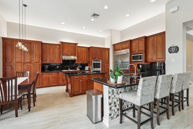 kitchen featuring decorative light fixtures, built in appliances, decorative backsplash, a kitchen bar, and a center island with sink