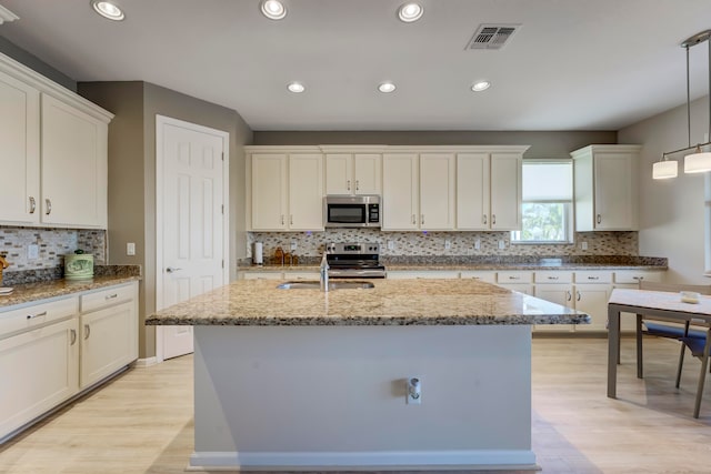 kitchen with tasteful backsplash, light wood-type flooring, an island with sink, stainless steel appliances, and decorative light fixtures