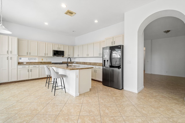 kitchen with light tile patterned flooring, sink, dark stone countertops, refrigerator with ice dispenser, and a kitchen island with sink