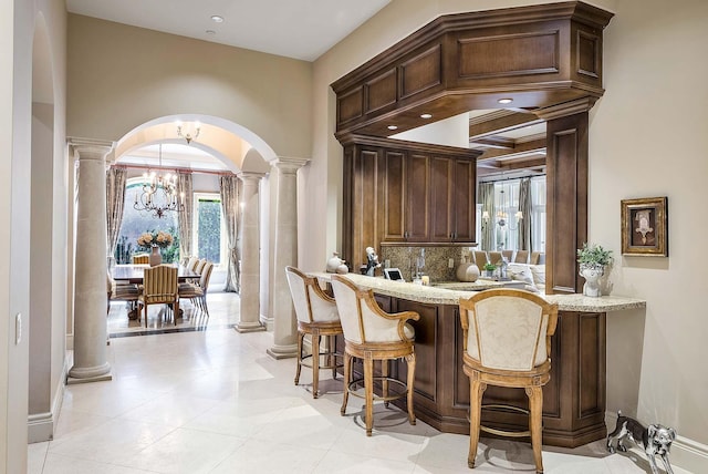 bar with dark brown cabinetry, a chandelier, light stone countertops, ornate columns, and decorative backsplash