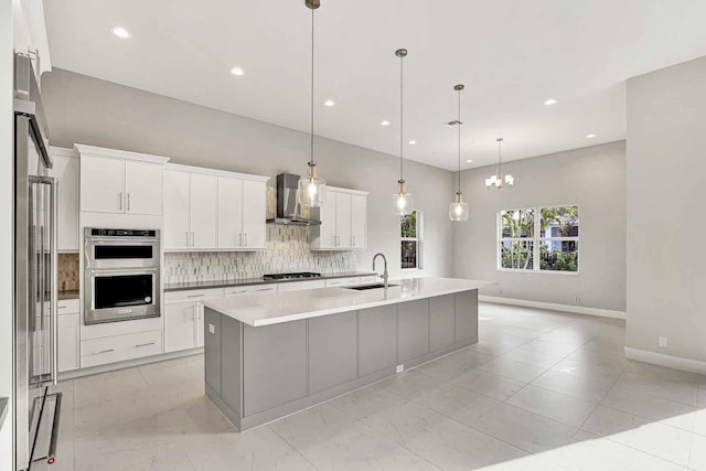 kitchen featuring pendant lighting, wall chimney range hood, appliances with stainless steel finishes, white cabinets, and a center island with sink