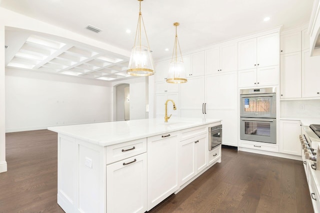 kitchen with white cabinetry, sink, stainless steel double oven, hanging light fixtures, and a center island with sink