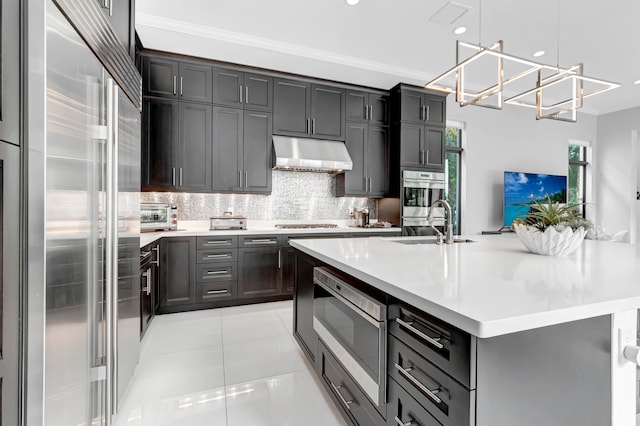 kitchen with built in appliances, an inviting chandelier, light tile patterned floors, backsplash, and a kitchen island with sink