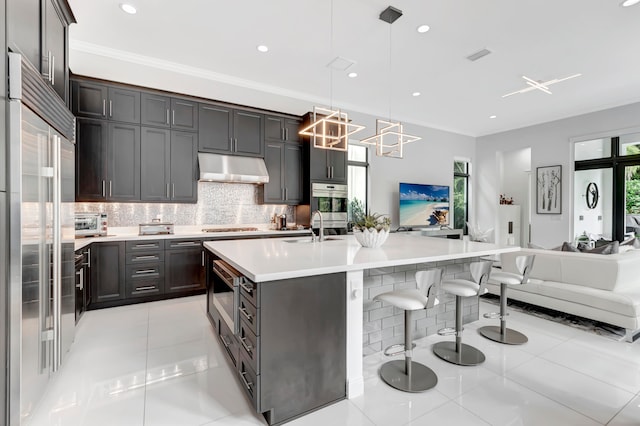 kitchen featuring built in appliances, backsplash, light tile patterned floors, dark brown cabinetry, and a kitchen island with sink