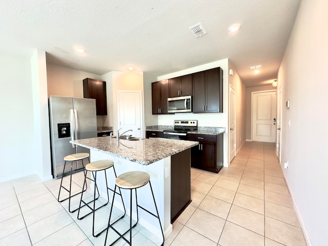 kitchen featuring dark brown cabinets, light tile patterned floors, an island with sink, stainless steel appliances, and a kitchen bar