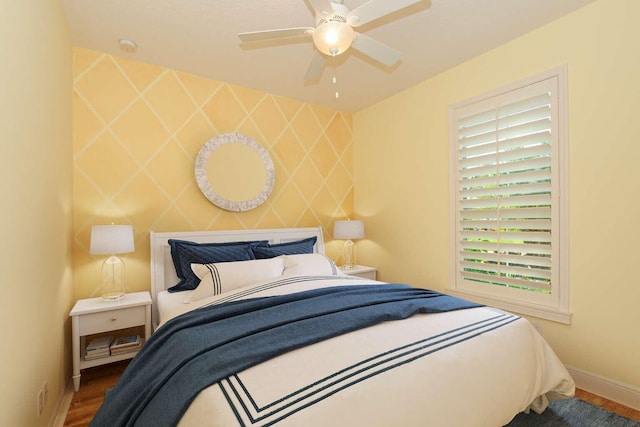 bedroom with ceiling fan, an accent wall, baseboards, and wood finished floors