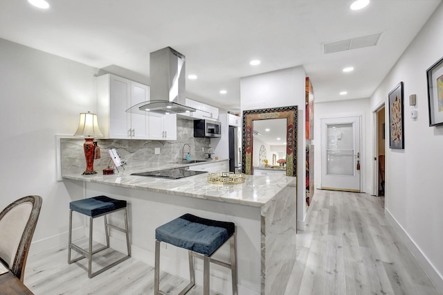 kitchen with light wood-type flooring, island exhaust hood, kitchen peninsula, white cabinets, and light stone counters