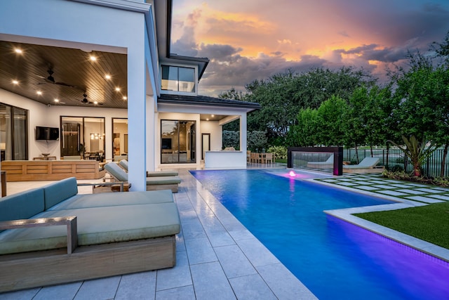 pool at dusk with a patio, outdoor lounge area, ceiling fan, and pool water feature