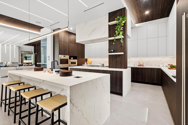 kitchen featuring dark brown cabinetry, light stone counters, tasteful backsplash, a center island with sink, and a kitchen breakfast bar