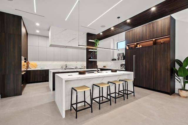 kitchen with a center island with sink, dark brown cabinetry, decorative backsplash, and a breakfast bar