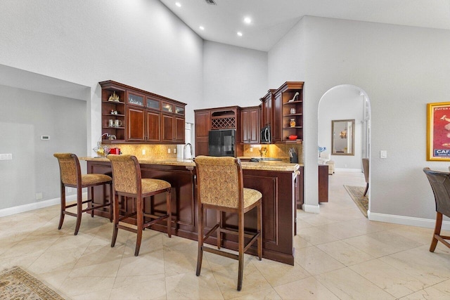 kitchen featuring a towering ceiling, a kitchen bar, backsplash, light stone counters, and fridge