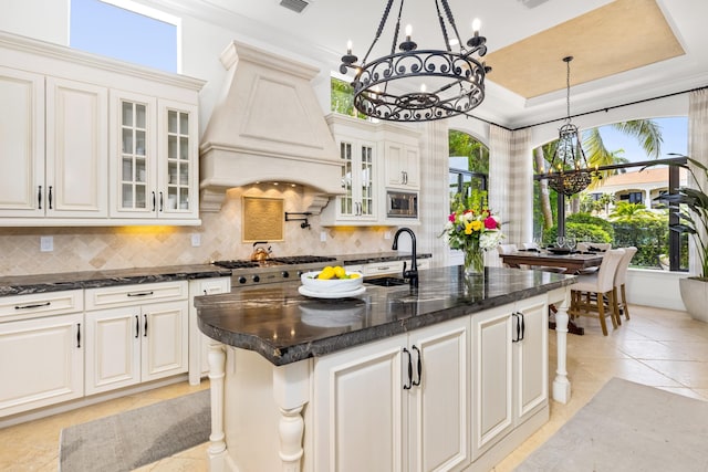 kitchen featuring sink, backsplash, an island with sink, light tile patterned flooring, and custom range hood