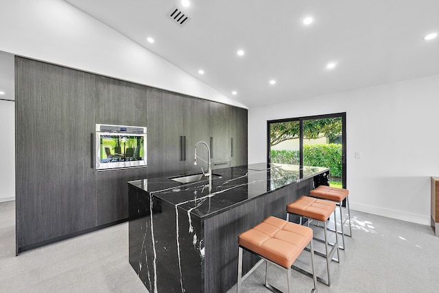 kitchen with a kitchen bar, stainless steel oven, a kitchen island with sink, dark stone counters, and sink