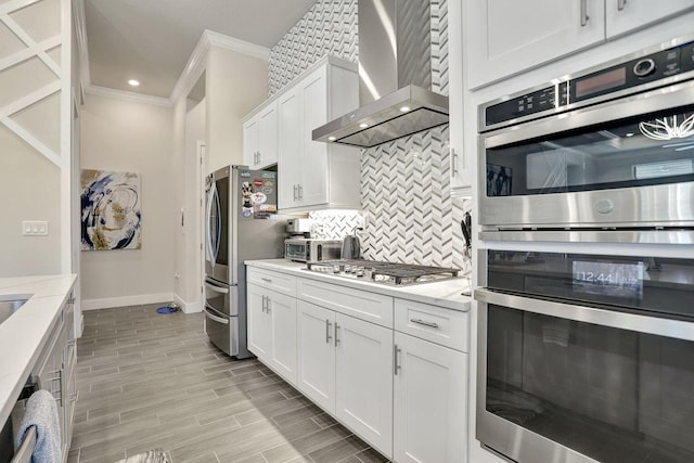 kitchen with appliances with stainless steel finishes, ornamental molding, white cabinets, decorative backsplash, and wall chimney exhaust hood