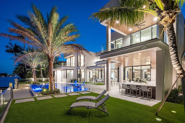 back house at night with a lawn, a patio area, a balcony, and exterior bar