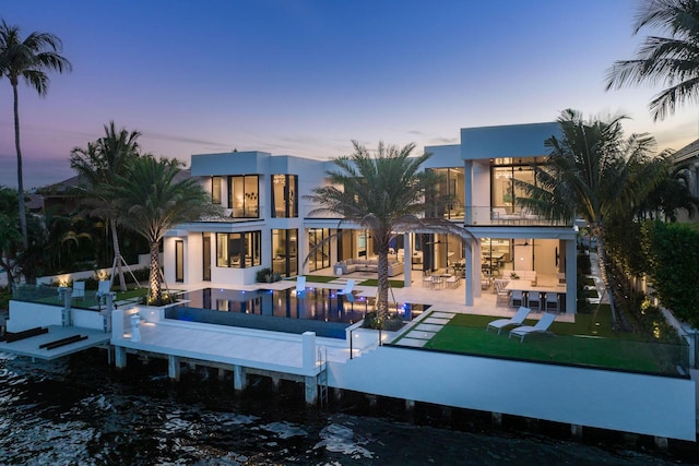 back house at dusk featuring a water view, a balcony, a patio, and a lawn