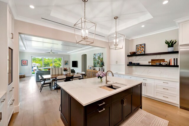 kitchen featuring light wood-type flooring, a kitchen island with sink, decorative light fixtures, a raised ceiling, and sink