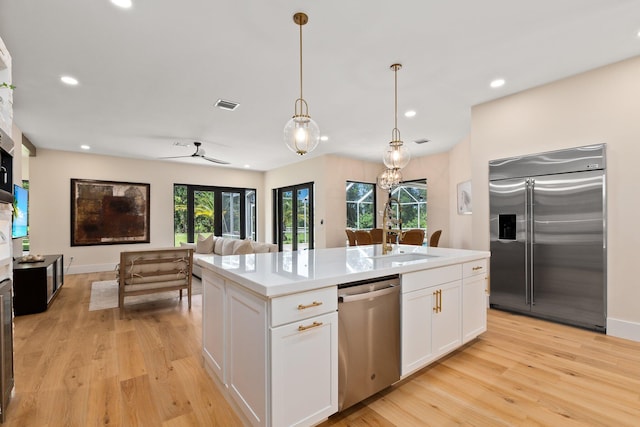 kitchen with appliances with stainless steel finishes, a center island with sink, pendant lighting, light wood-type flooring, and white cabinetry