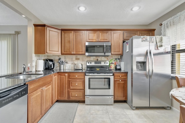 kitchen with light tile patterned flooring, a sink, appliances with stainless steel finishes, brown cabinetry, and dark stone countertops