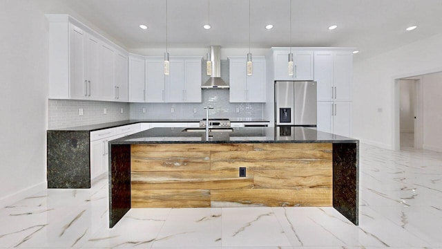 kitchen featuring stainless steel fridge, backsplash, an island with sink, light tile patterned flooring, and wall chimney exhaust hood