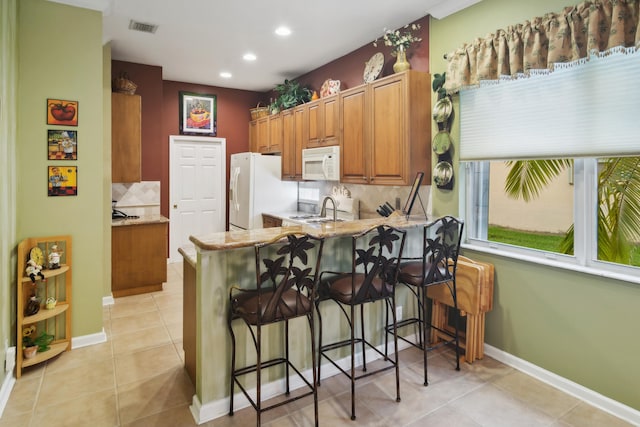 kitchen with light tile patterned floors, a kitchen breakfast bar, kitchen peninsula, white appliances, and backsplash