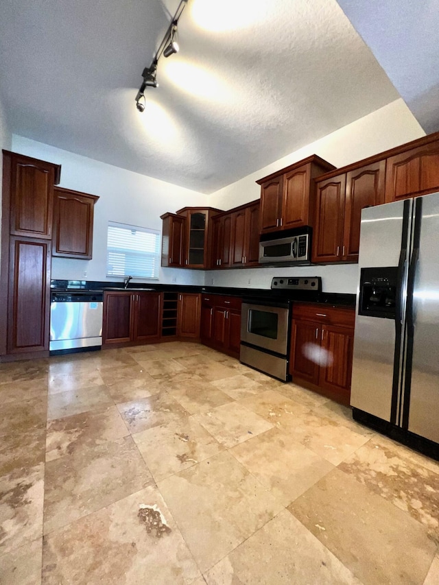 kitchen with a textured ceiling, a sink, appliances with stainless steel finishes, dark countertops, and glass insert cabinets