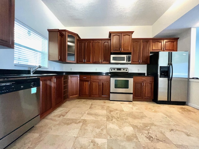 kitchen with a textured ceiling, stainless steel appliances, a sink, dark countertops, and glass insert cabinets