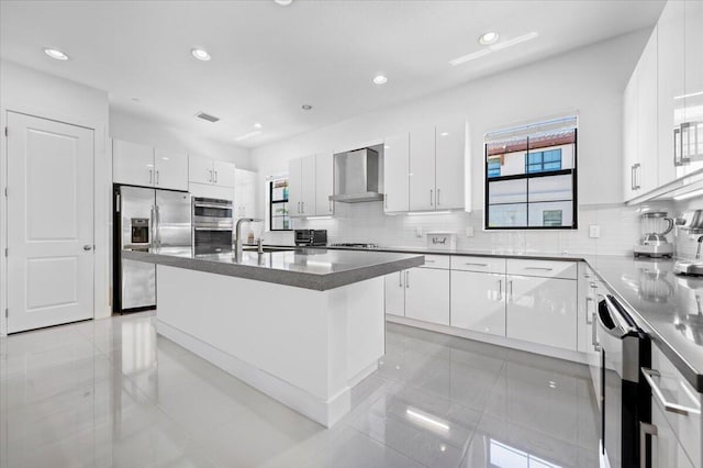 kitchen featuring a wealth of natural light, stainless steel appliances, wall chimney range hood, a center island with sink, and white cabinets