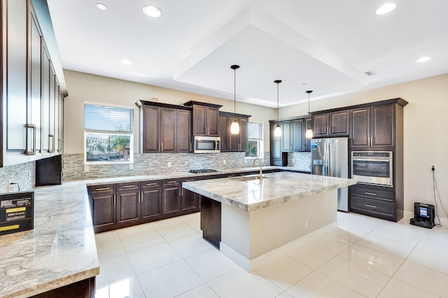 kitchen featuring sink, appliances with stainless steel finishes, pendant lighting, light stone countertops, and a kitchen island with sink