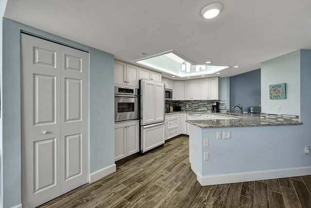 kitchen with appliances with stainless steel finishes, white cabinetry, backsplash, kitchen peninsula, and dark stone counters