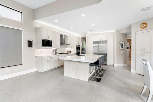 kitchen featuring wall chimney range hood, sink, a kitchen bar, built in appliances, and a kitchen island with sink