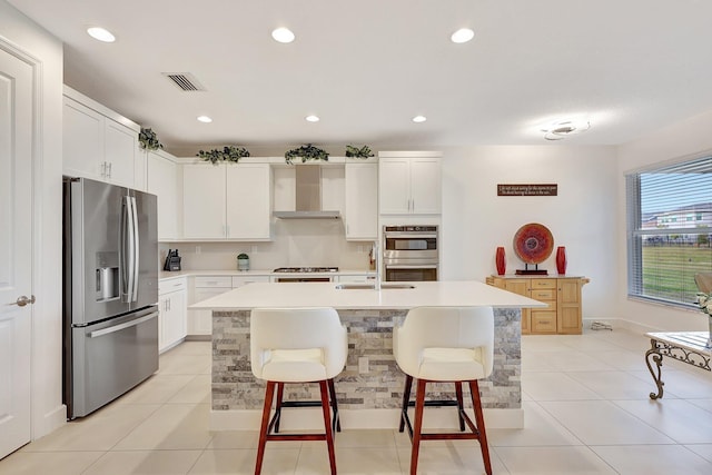 kitchen with a kitchen bar, white cabinetry, wall chimney range hood, stainless steel appliances, and a center island with sink