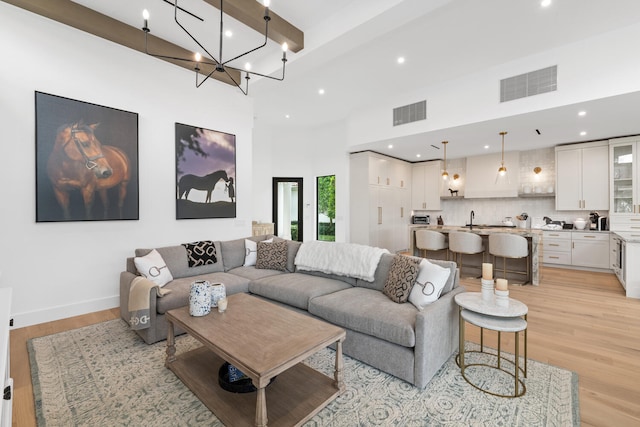 living room featuring light hardwood / wood-style flooring, a chandelier, beamed ceiling, a towering ceiling, and sink