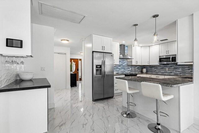 kitchen featuring stainless steel appliances, white cabinetry, wall chimney range hood, decorative light fixtures, and tasteful backsplash